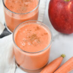 Two apple carrot smoothies next to an apple and peeled carrots on a table