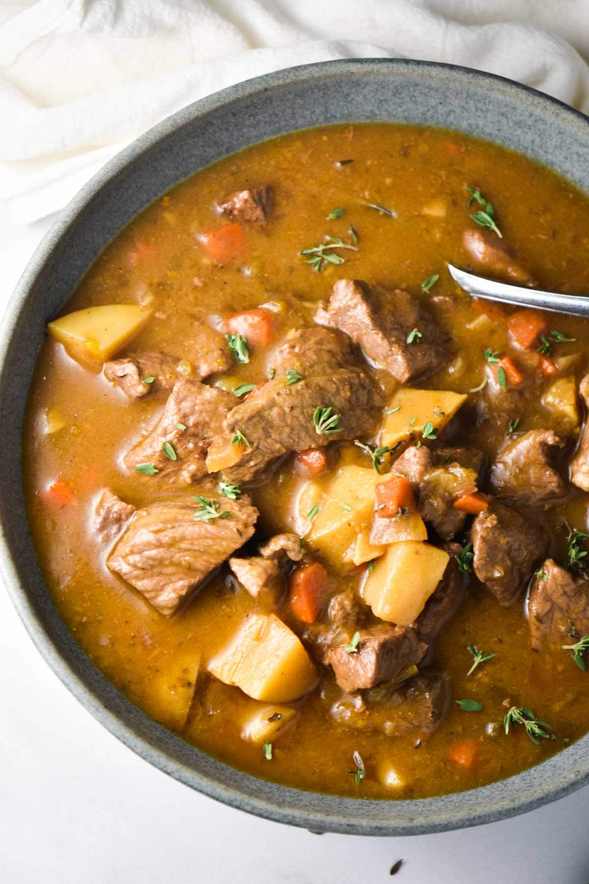 A close up of beef chuck roast, potatoes, carrots, and thyme in a stew with a spoon dipping in