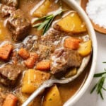A spoon scooping a thick beef stew with potatoes, carrots, and celery, topped with a rosemary leaf.