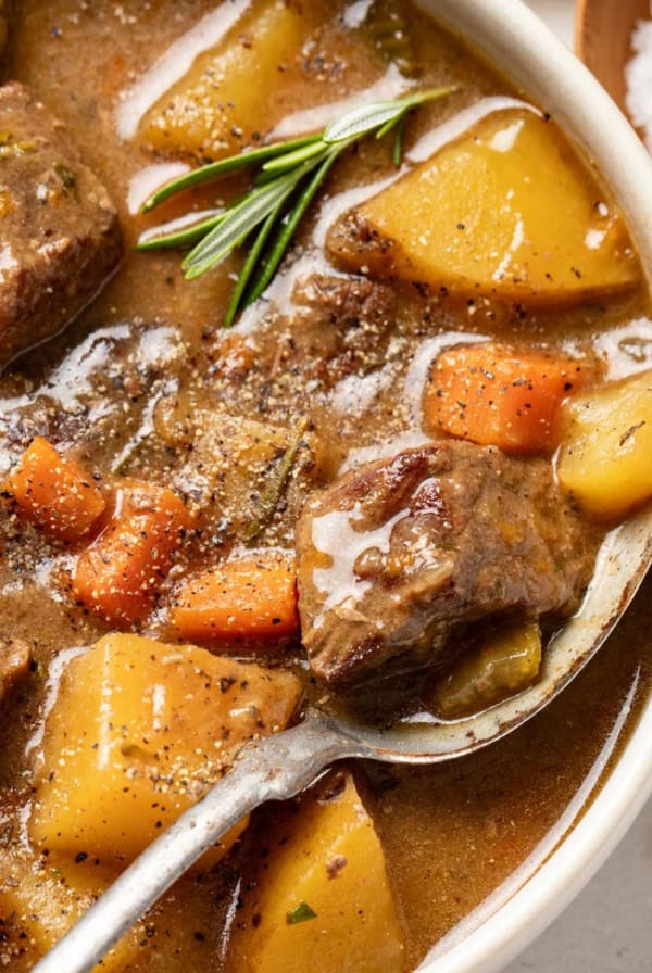 A spoon scooping a thick beef stew with potatoes, carrots, and celery, topped with a rosemary leaf.