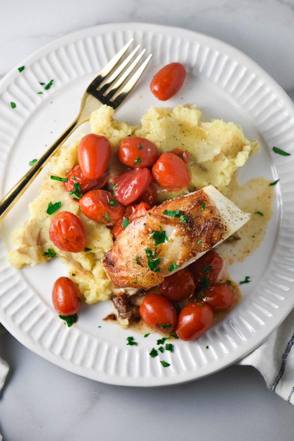 A white plate topped with mashed potatoes and a filet of seared sea bass topped with roasted tomatoes and a light sauce next to a gold fork. 