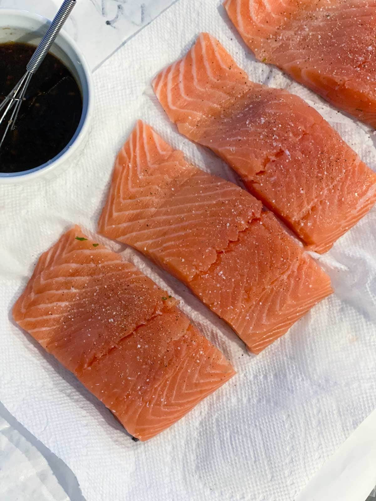 Four raw salmon fillets on a paper towel next to teriyaki sauce in a white bowl with a whisk.