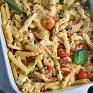 Baked goat cheese pasta in a white baking dish with tomatoes and basil.
