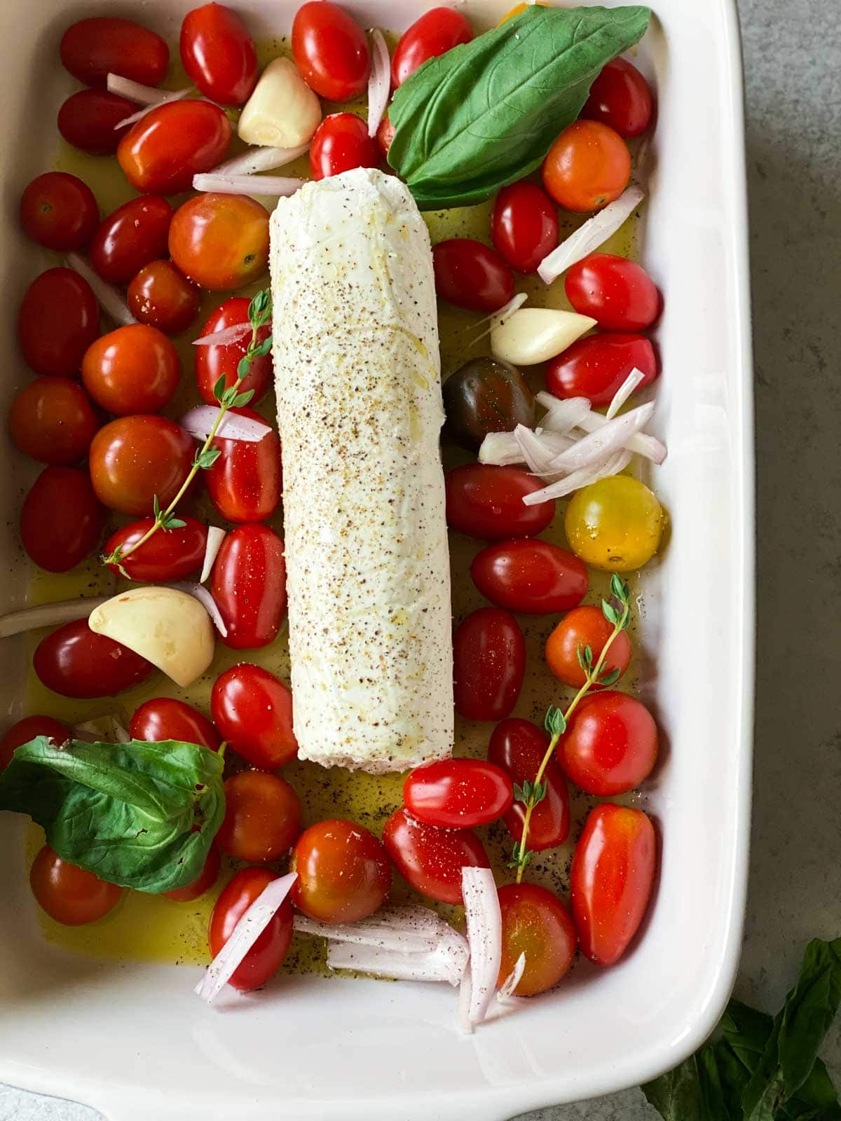 A log of goat cheese in a baking dish with tomatoes, fresh basil, garlic, and shallots.