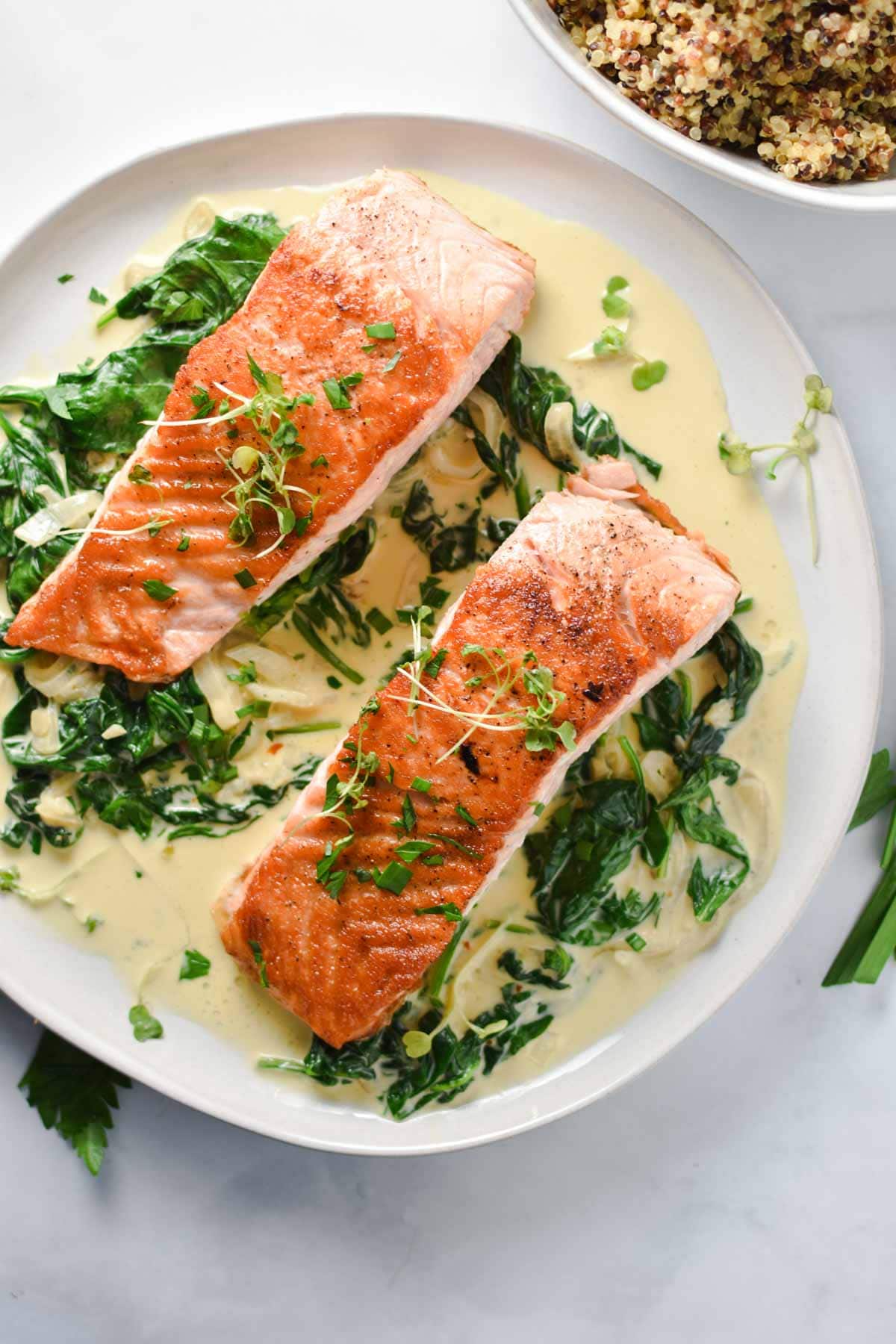 Two filets of salmon on top of spinach surrounded by a creamy sauce next to a bowl of quinoa.