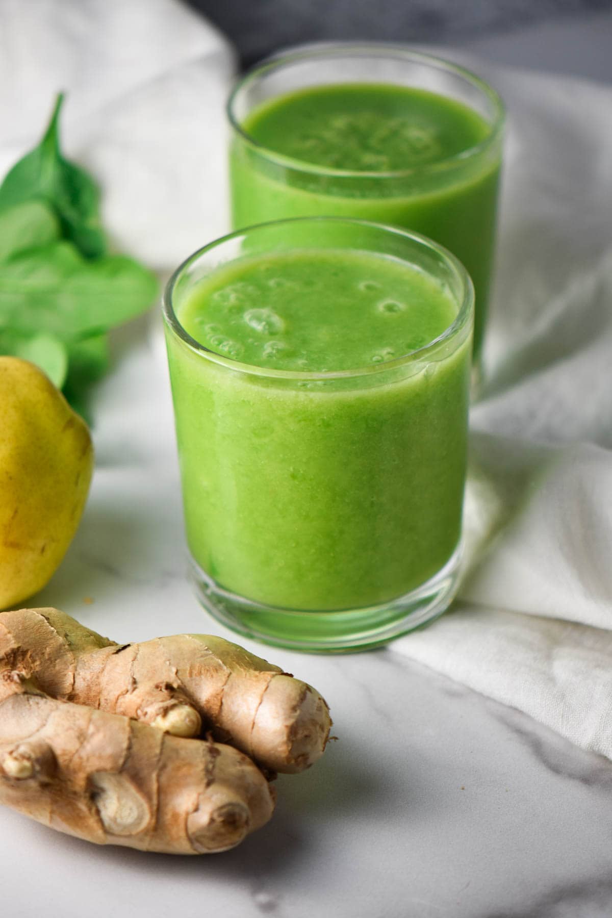 Two cups with a green anti inflammatory smoothie and ginger root on a white table.