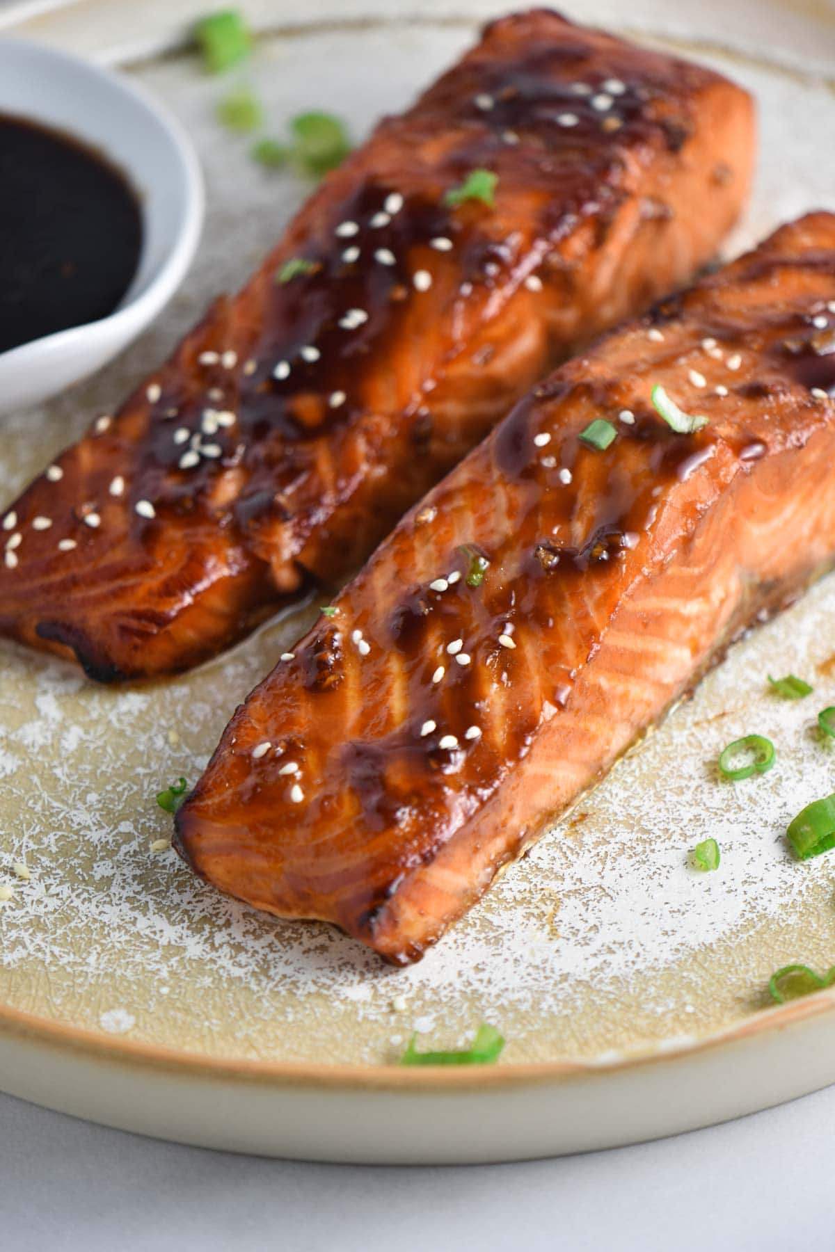 Two glazed air fryer salmon on a plate next to a bowl of teriyaki sauce