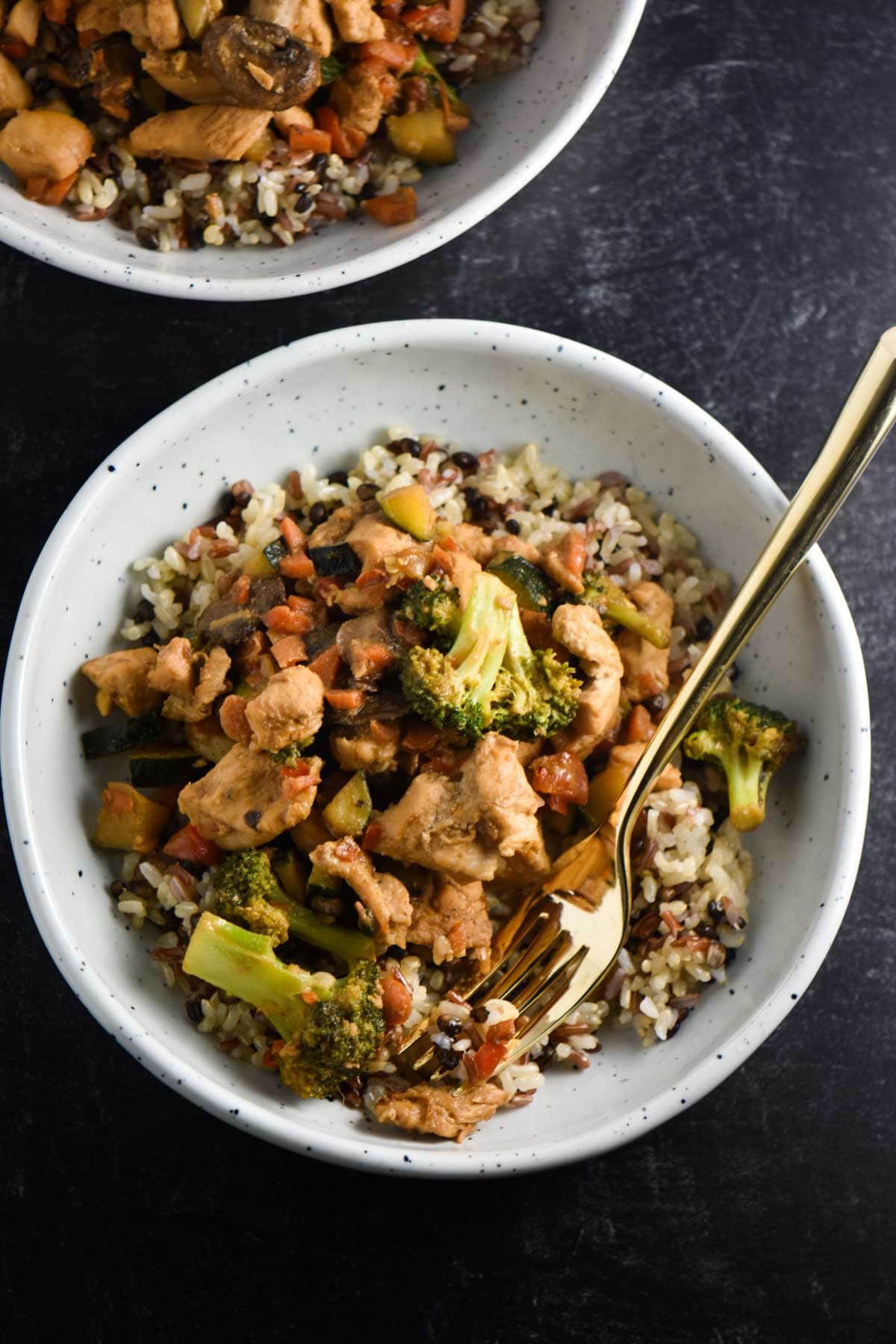 A bowl of soy free stir fry on a black table with a gold fork piercing a chicken piece.