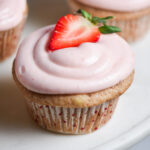 A pink strawberry cupcake with cream cheese frosting and a fresh strawberry on top.