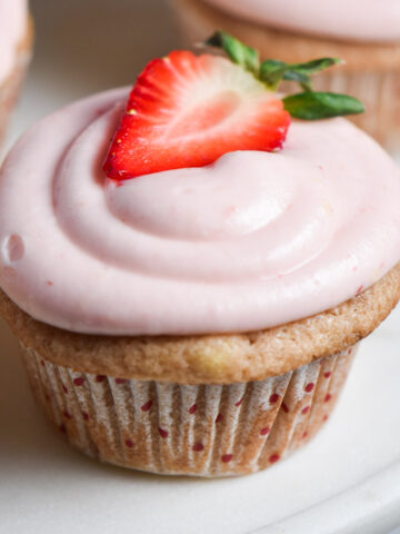 A pink strawberry cupcake with cream cheese frosting and a fresh strawberry on top.