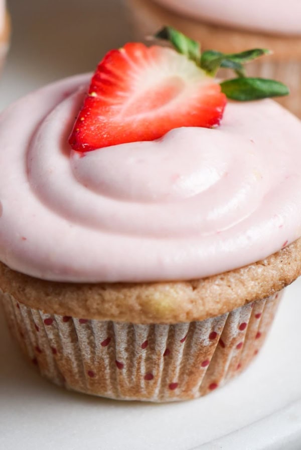 A pink strawberry cupcake with cream cheese frosting and a fresh strawberry on top.