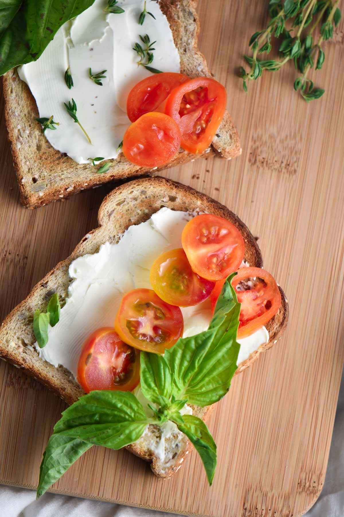 Toast with cream cheese, tomatoes, and basil on a wood board with fresh thyme.