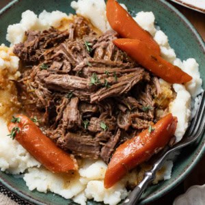 Shredded pot roast on top of creamy mashed potatoes with sauce and carrots in a green bowl.