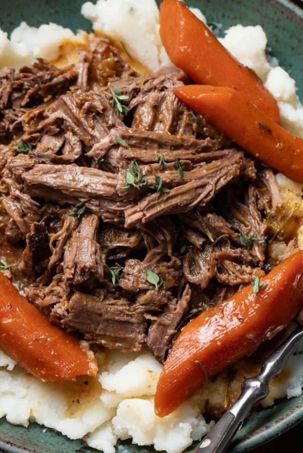 Shredded pot roast on top of creamy mashed potatoes with sauce and carrots in a green bowl.