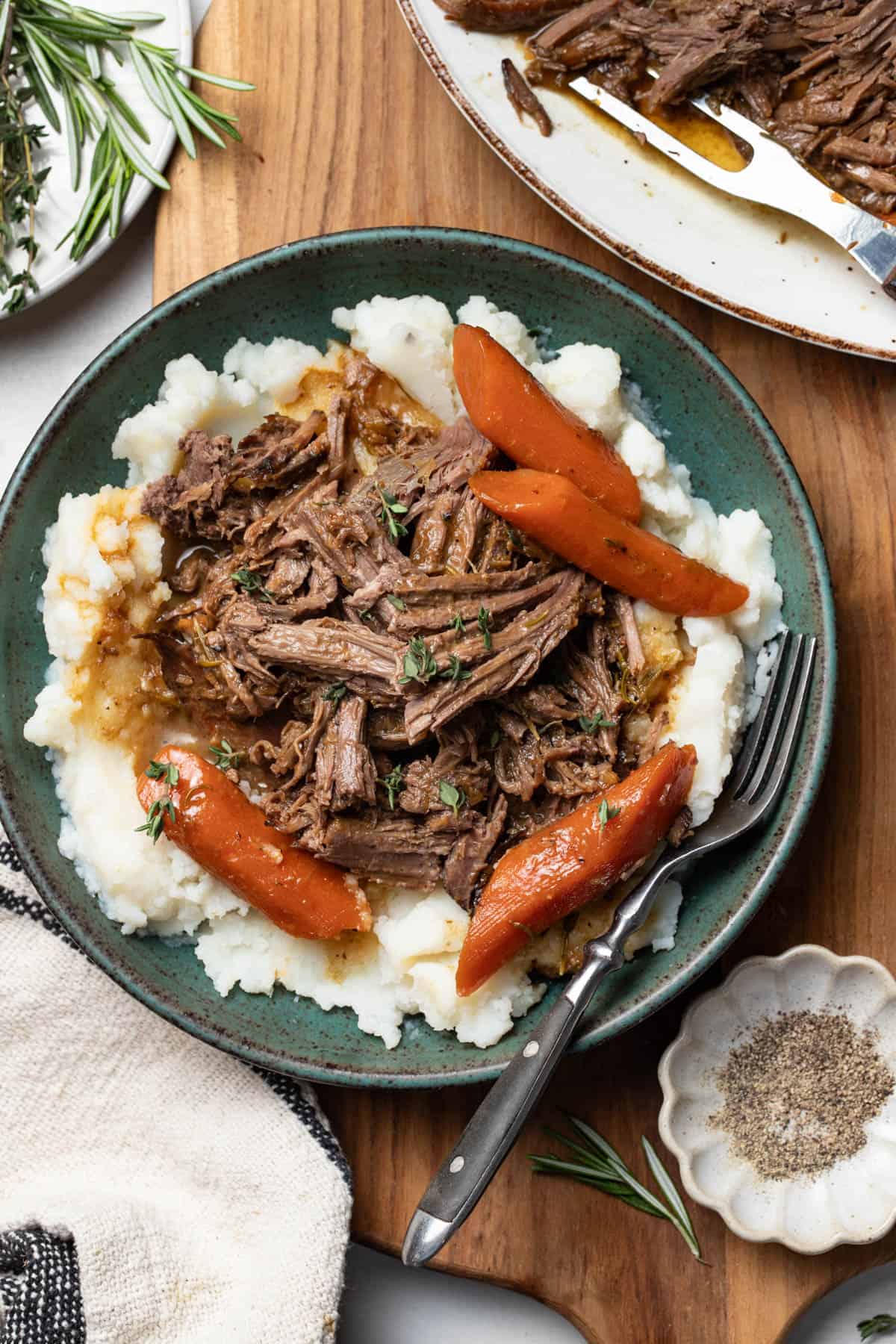 Classic pot roast spooned on top of mashed potatoes in a bowl on a cutting board with herbs.