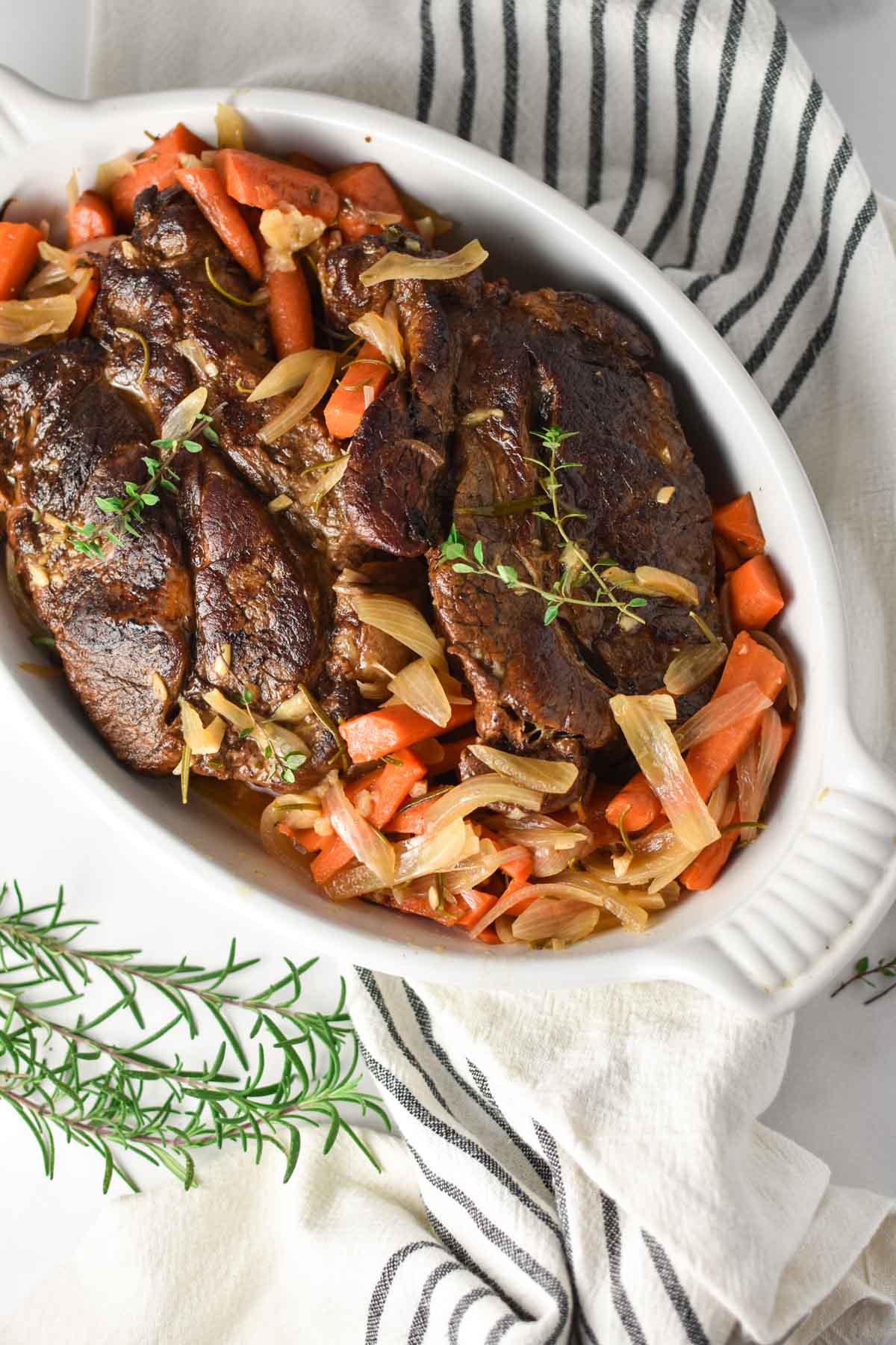 A beef chuck roast in a white baking dish with herbs.