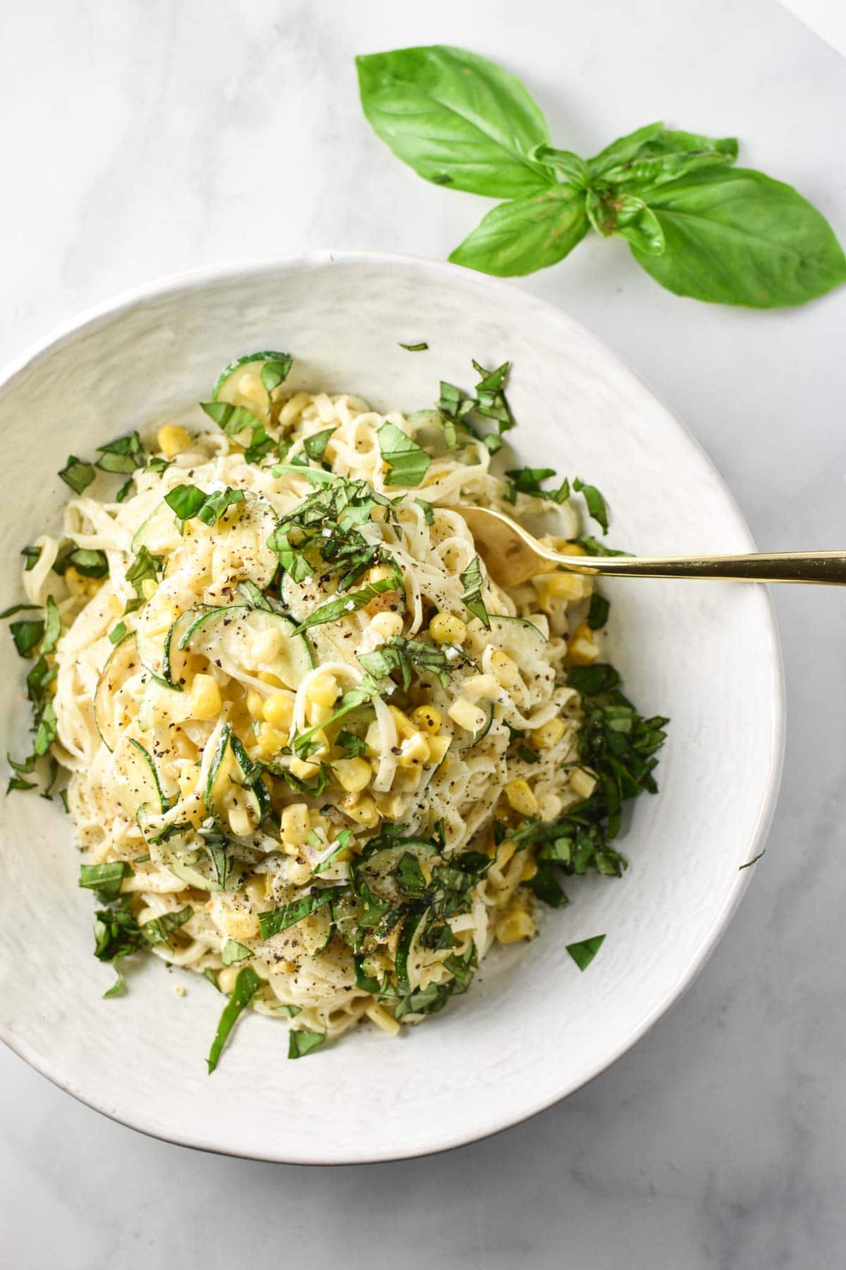 A fork digging into pasta with corn and zucchini, topped with basil and next to fresh basil leaves. 