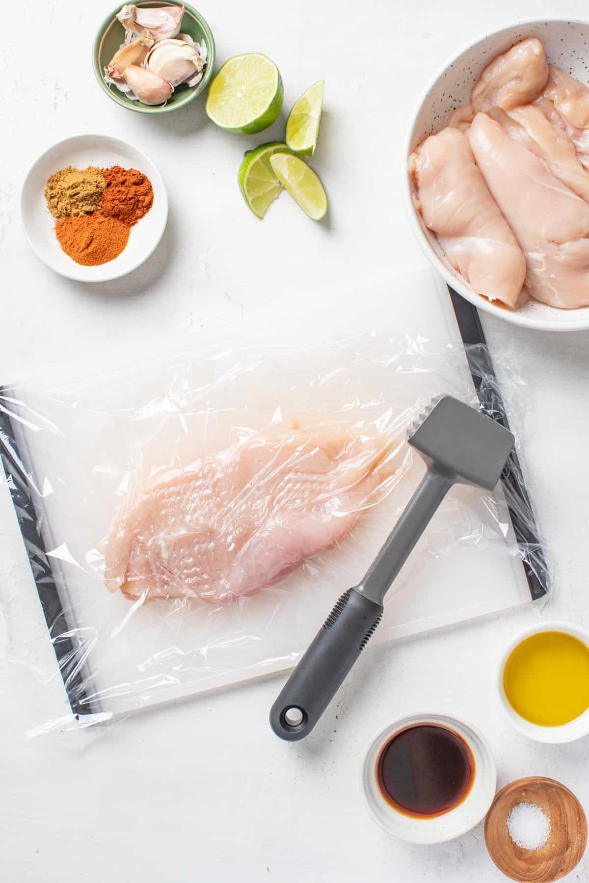 A cutting board topped with a raw chicken breast and a mallet, showing how to flatten the chicken breast before grilling. 