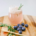 A blueberry mocktail on a wood table with blueberries and rosemary.