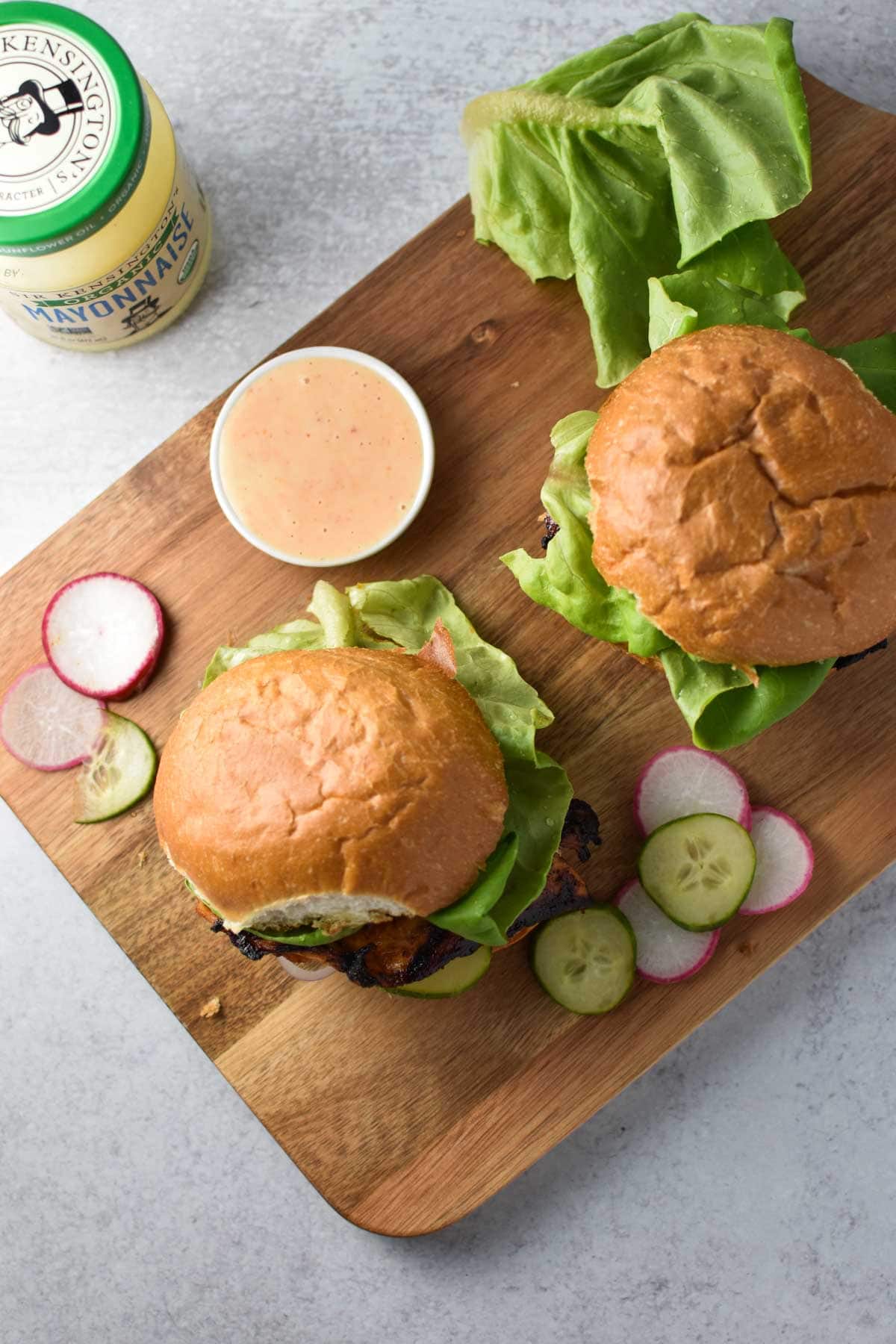 Two grilled chicken sandwiches on a board with cucumbers and spicy mayo.