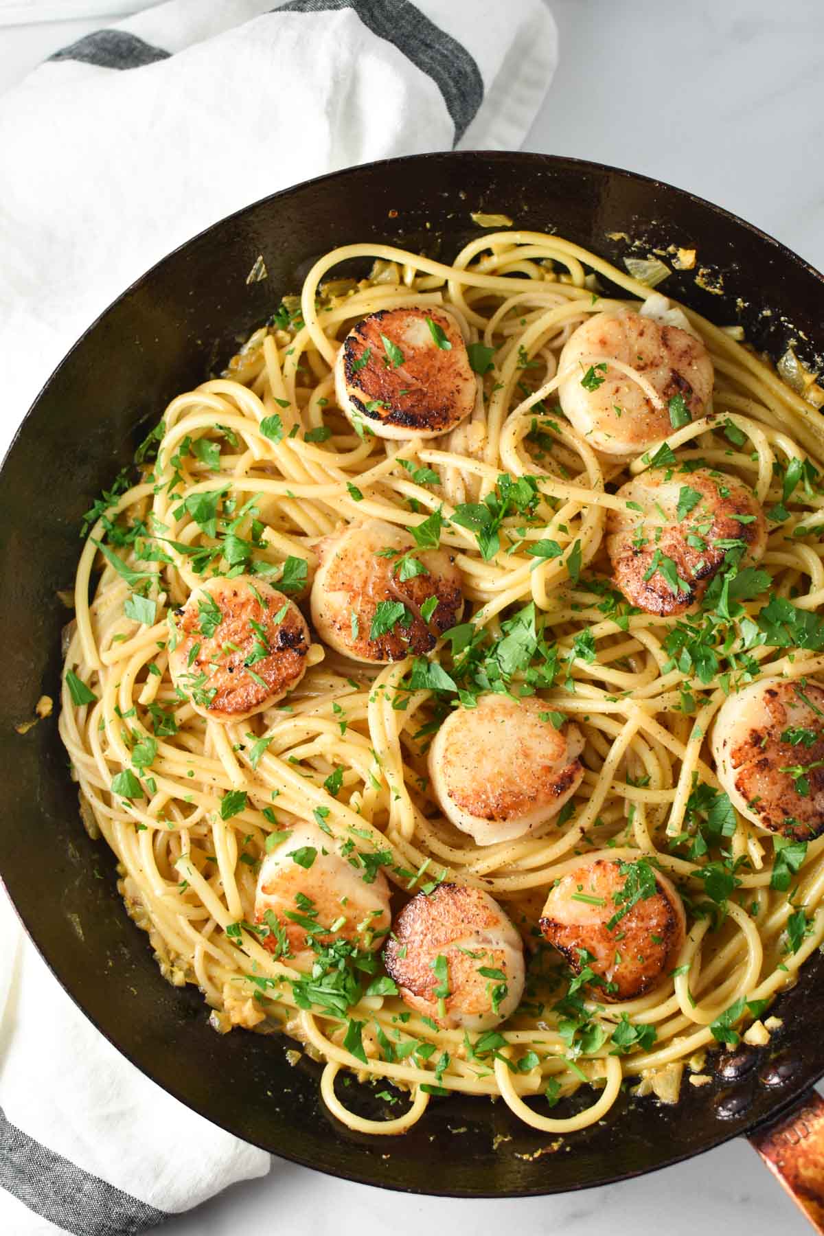 Pasta topped with seared scallops in a black pan with a towel.