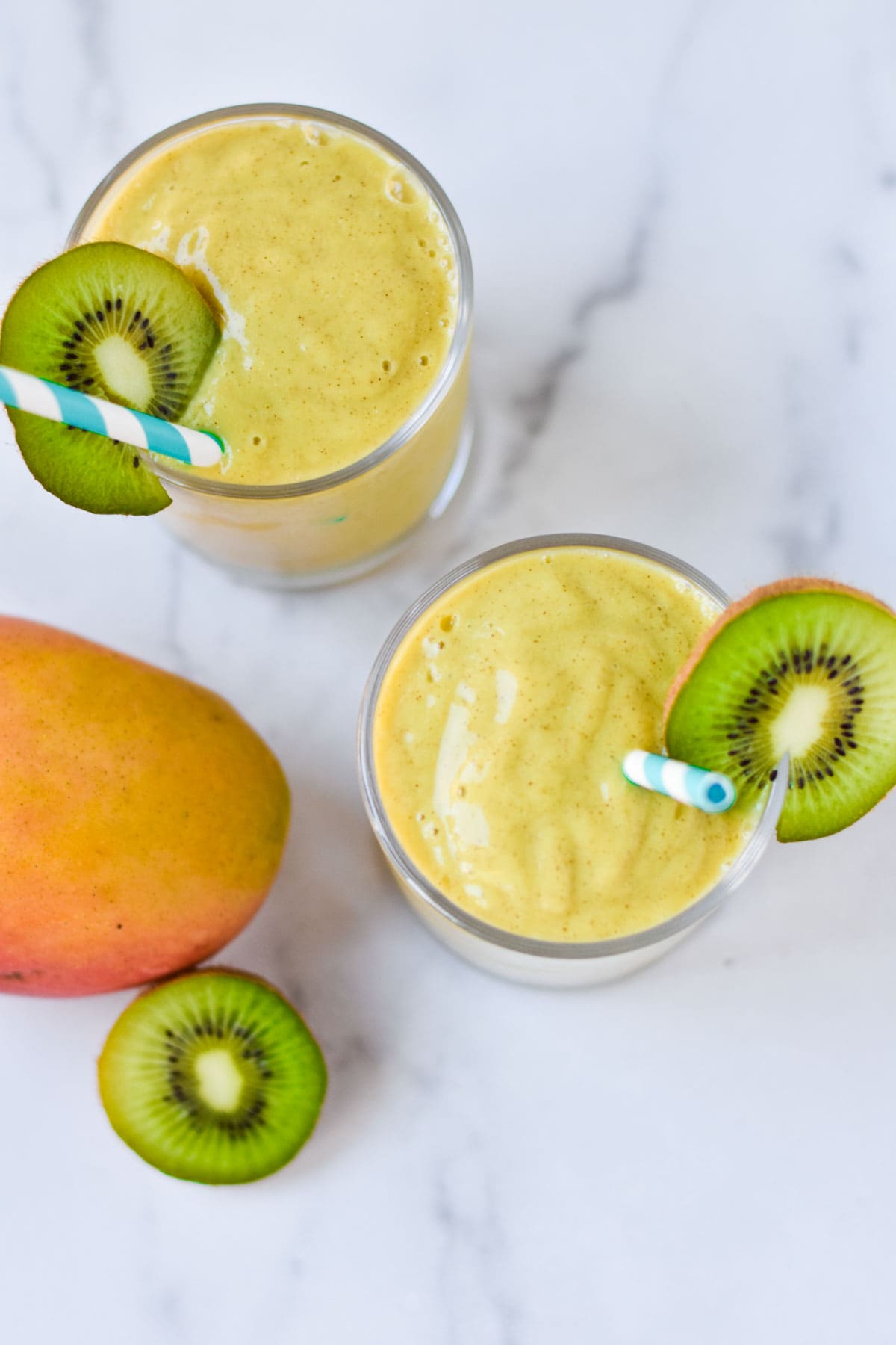 Two glasses of a bright yellow smoothie next to a fresh mango and kiwi fruit.