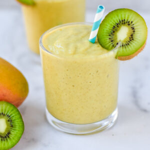A mango smoothie in a clear glass with a kiwi and blue striped straw.