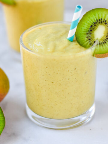 A mango smoothie in a clear glass with a kiwi and blue striped straw.