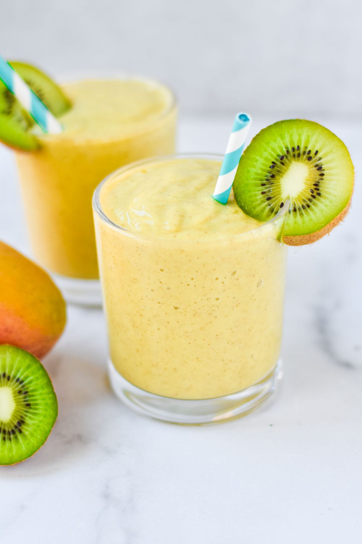 Two kiwi and mango smoothies in short glasses on a marble surface.