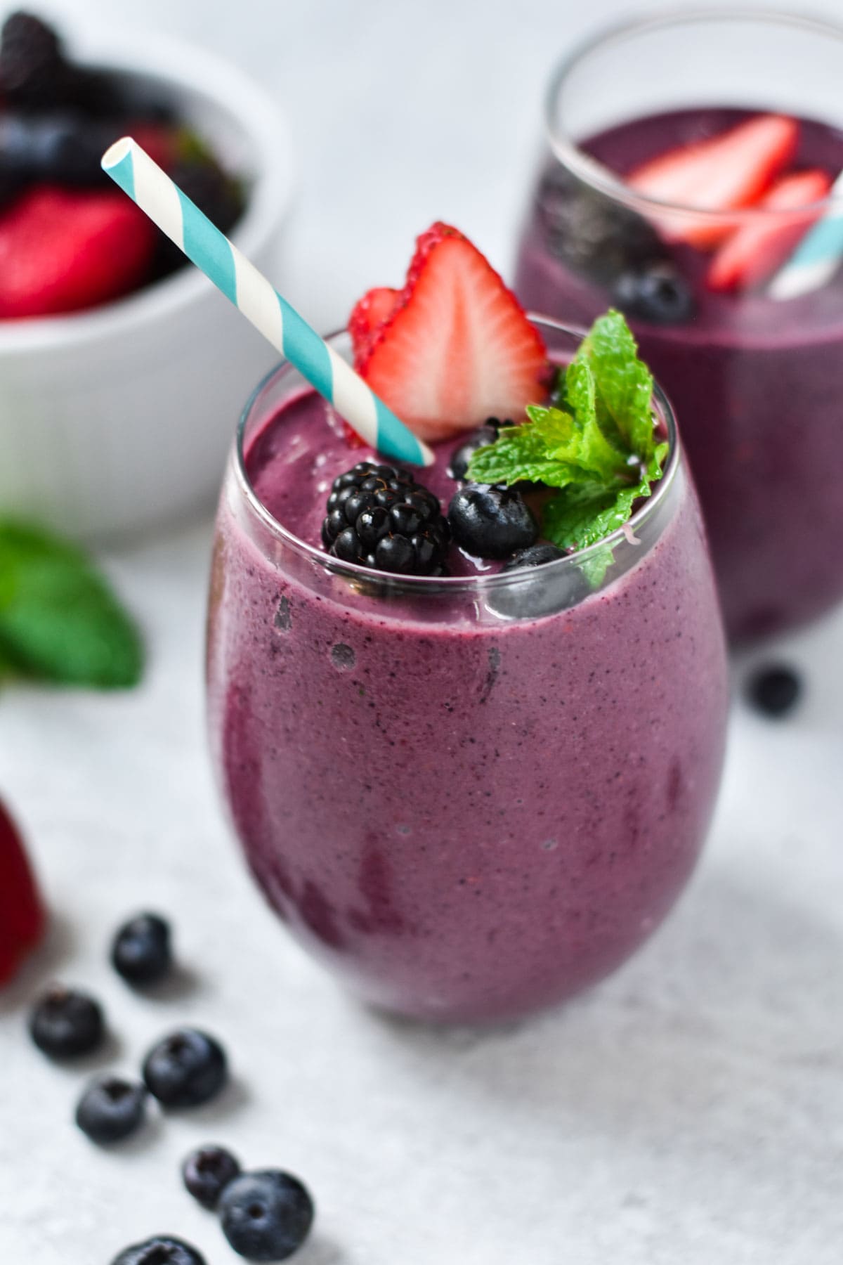 A purple mixed berry smoothie with mint, strawberries, and a straw in a glass. 
