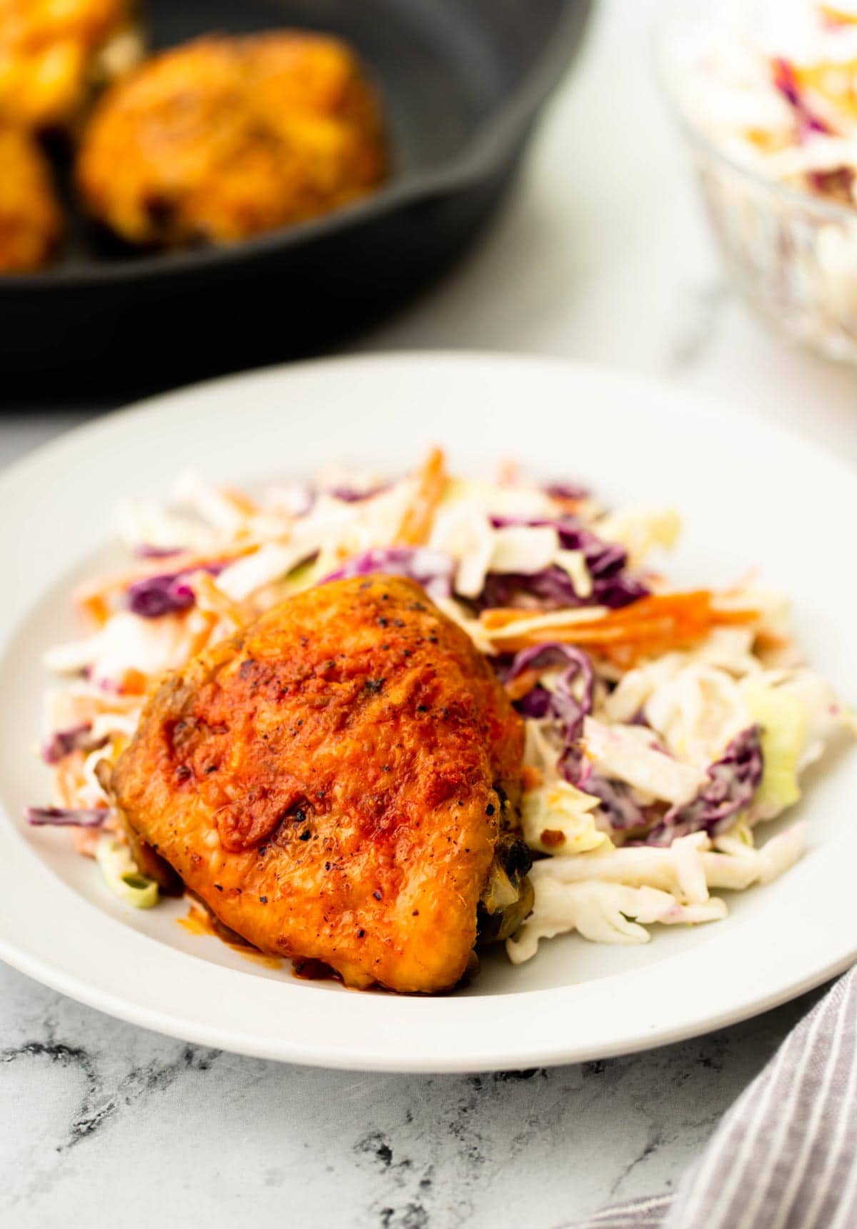 Crispy chicken thighs on a white plate with coleslaw and a cast iron pan in the back.