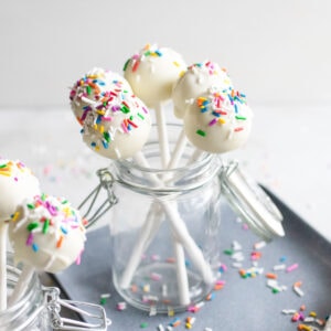 Cake pops with bright colored sprinkles in a glass jar on a baking sheet.