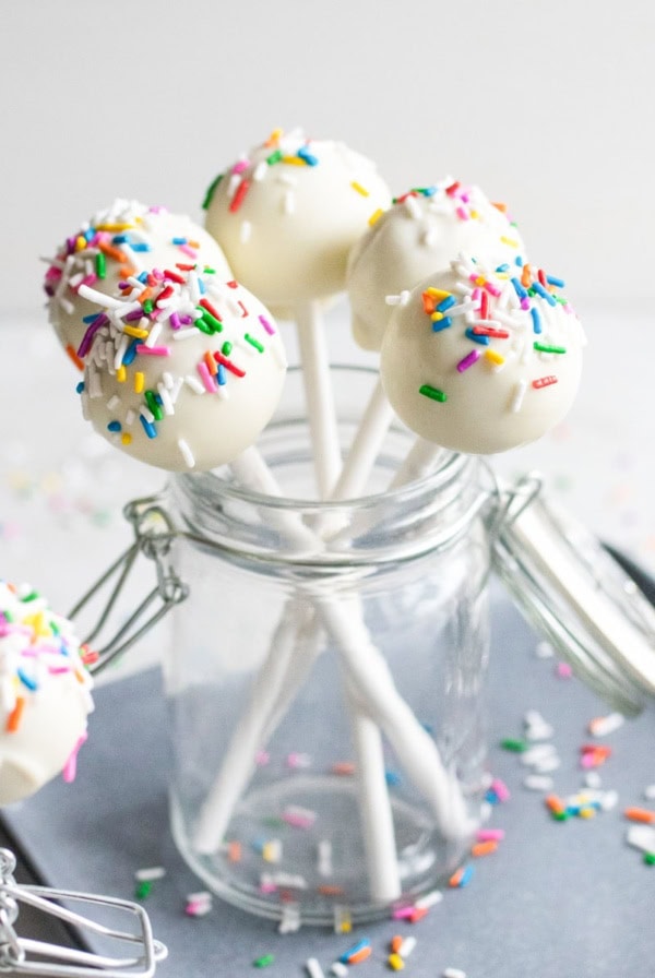 Cake pops with bright colored sprinkles in a glass jar on a baking sheet.