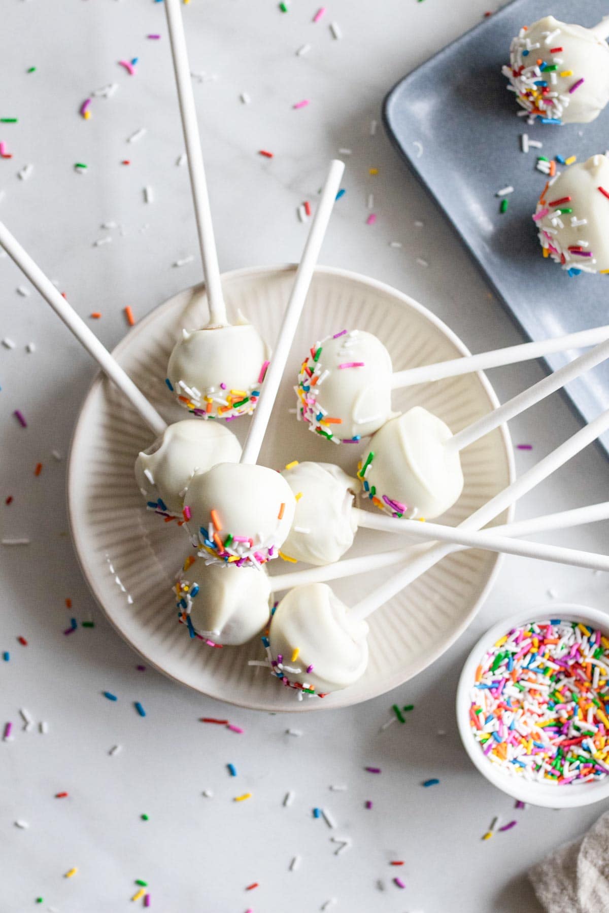 Cake pops stacked on top of each other on a plate next to colorful sprinkles. 