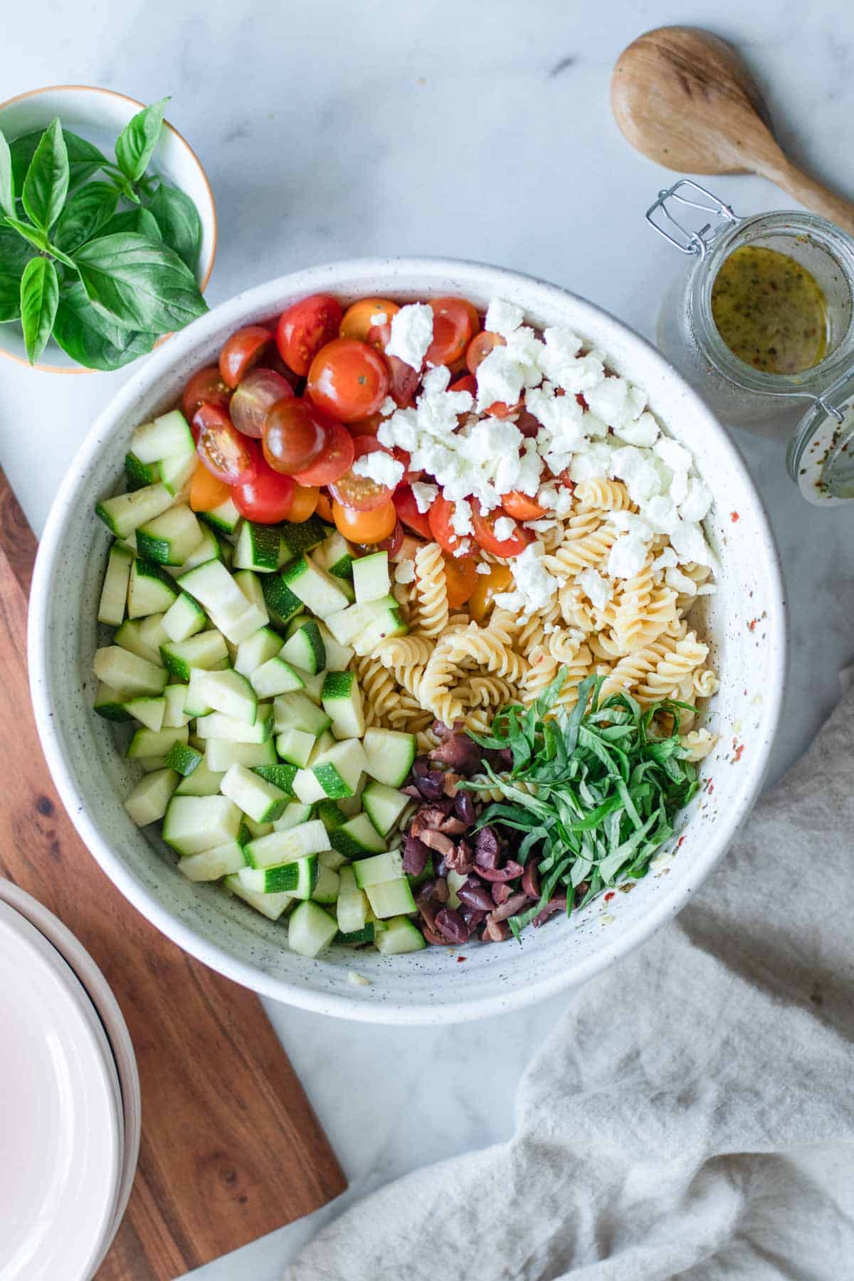 A bowl of the ingredients for pasta salad all separated next to wooden spoons.