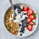 A spoon dipped into cottage cheese with fruit, granola, and seeds.