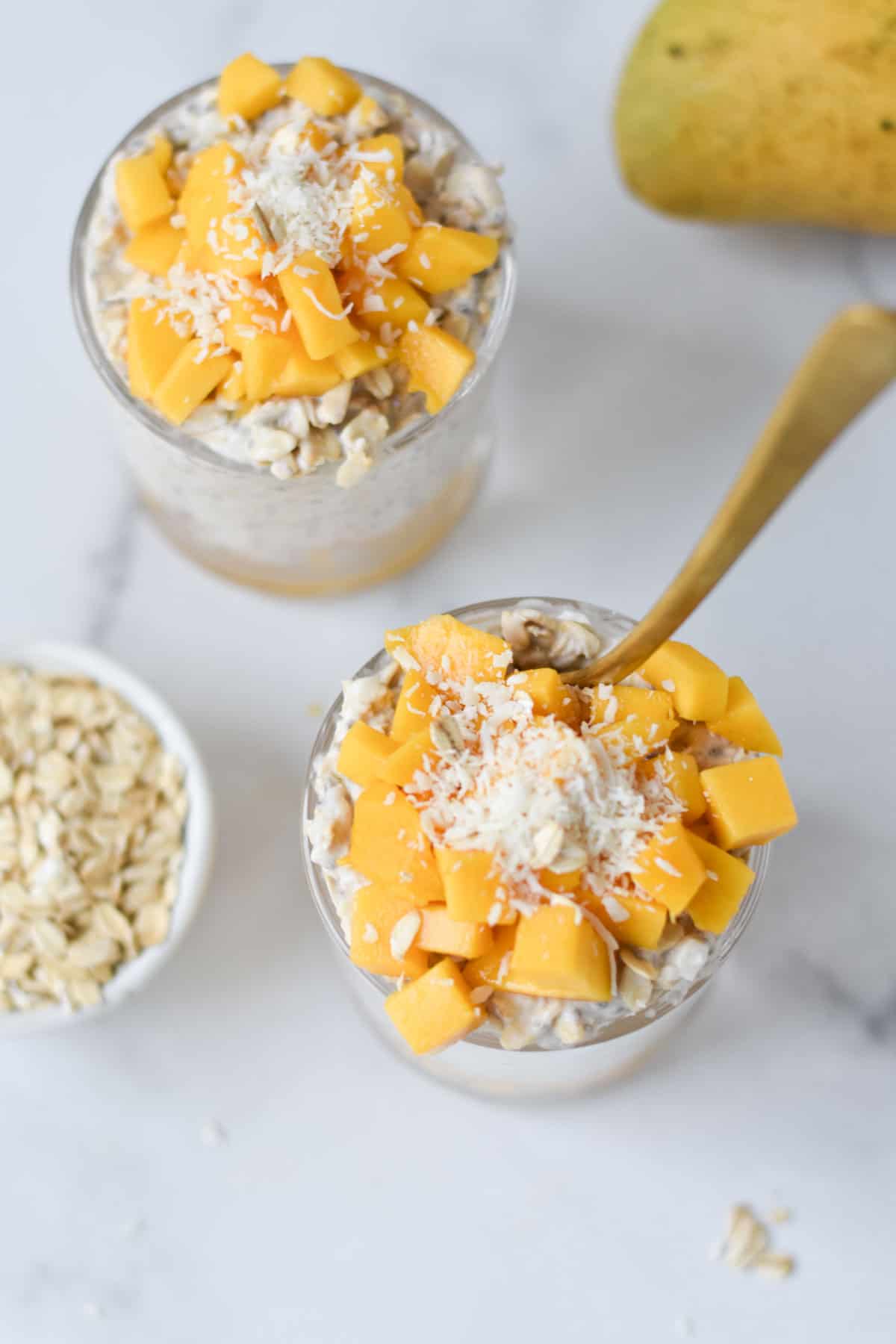 Two cups of mango overnight oats with spoons and coconut on top.