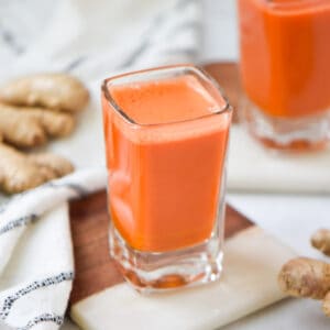 Two carrot wellness shots next to fresh ginger root.