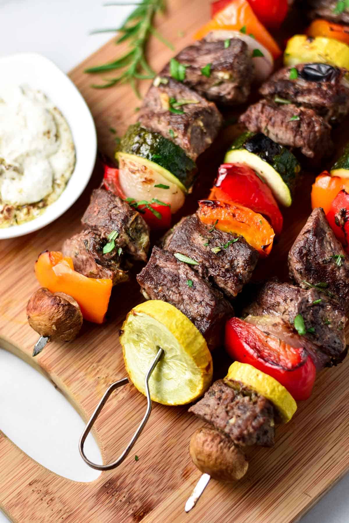 Three beef kabobs on a wood board next to a dip and rosemary.