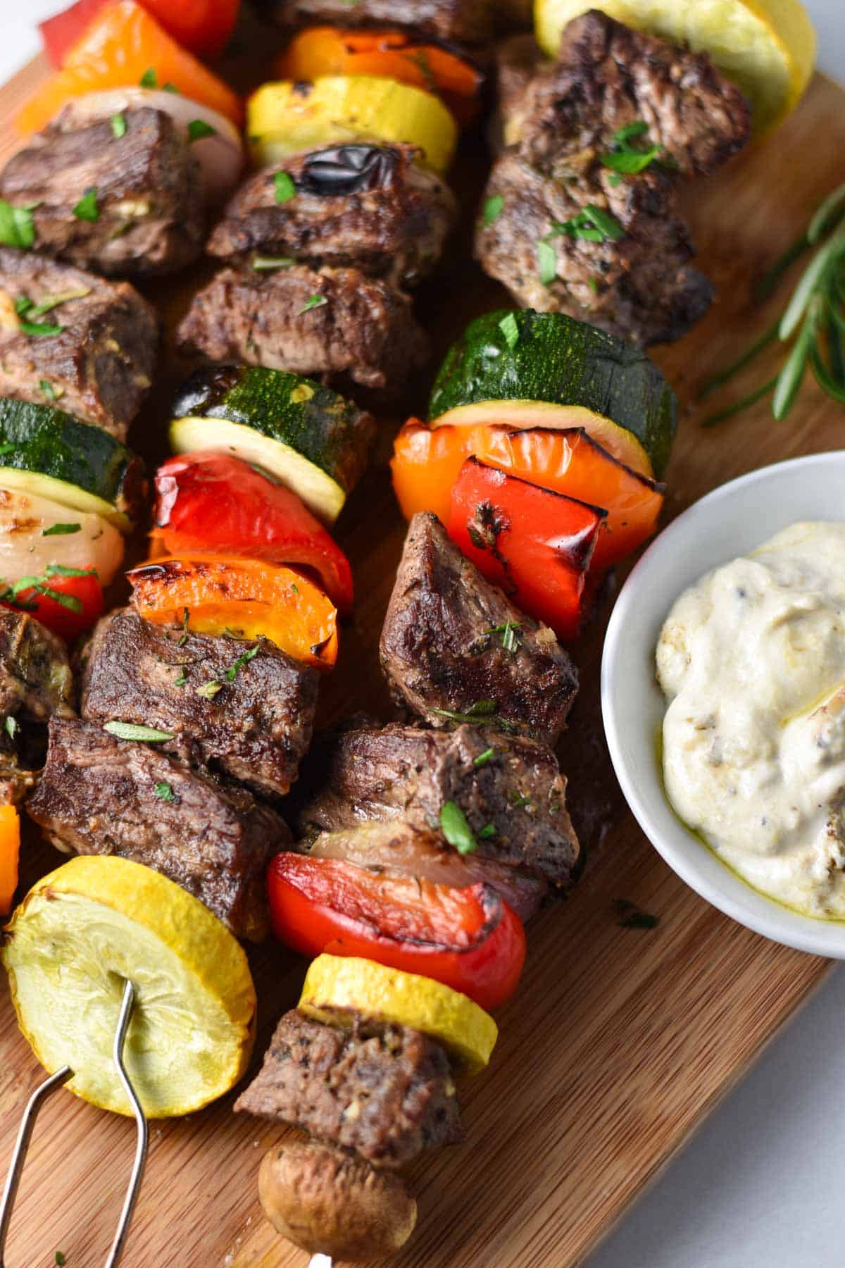Three beef and vegetable skewers on a wood board next to a dip and rosemary.