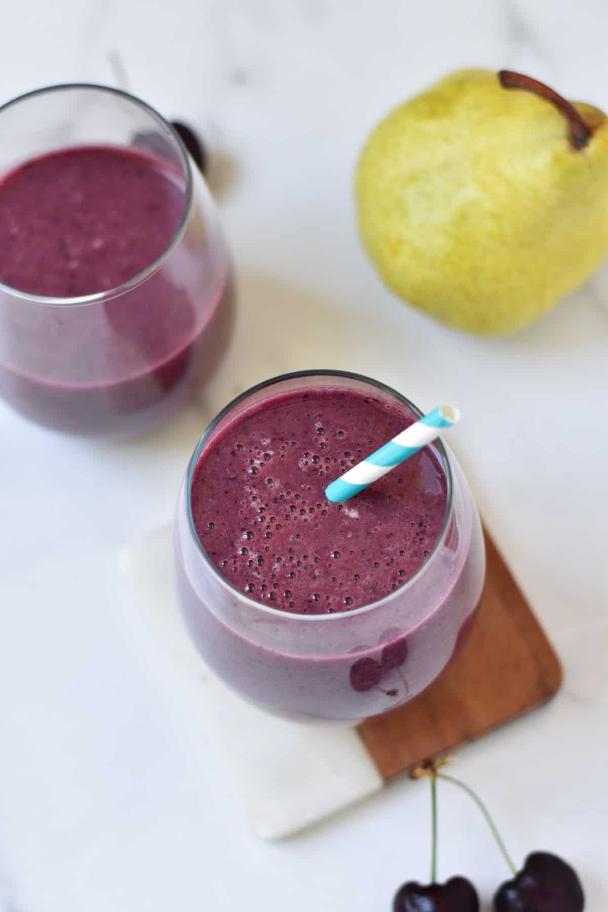 Two berry smoothies in glass cups next to a whole pear and cherries.