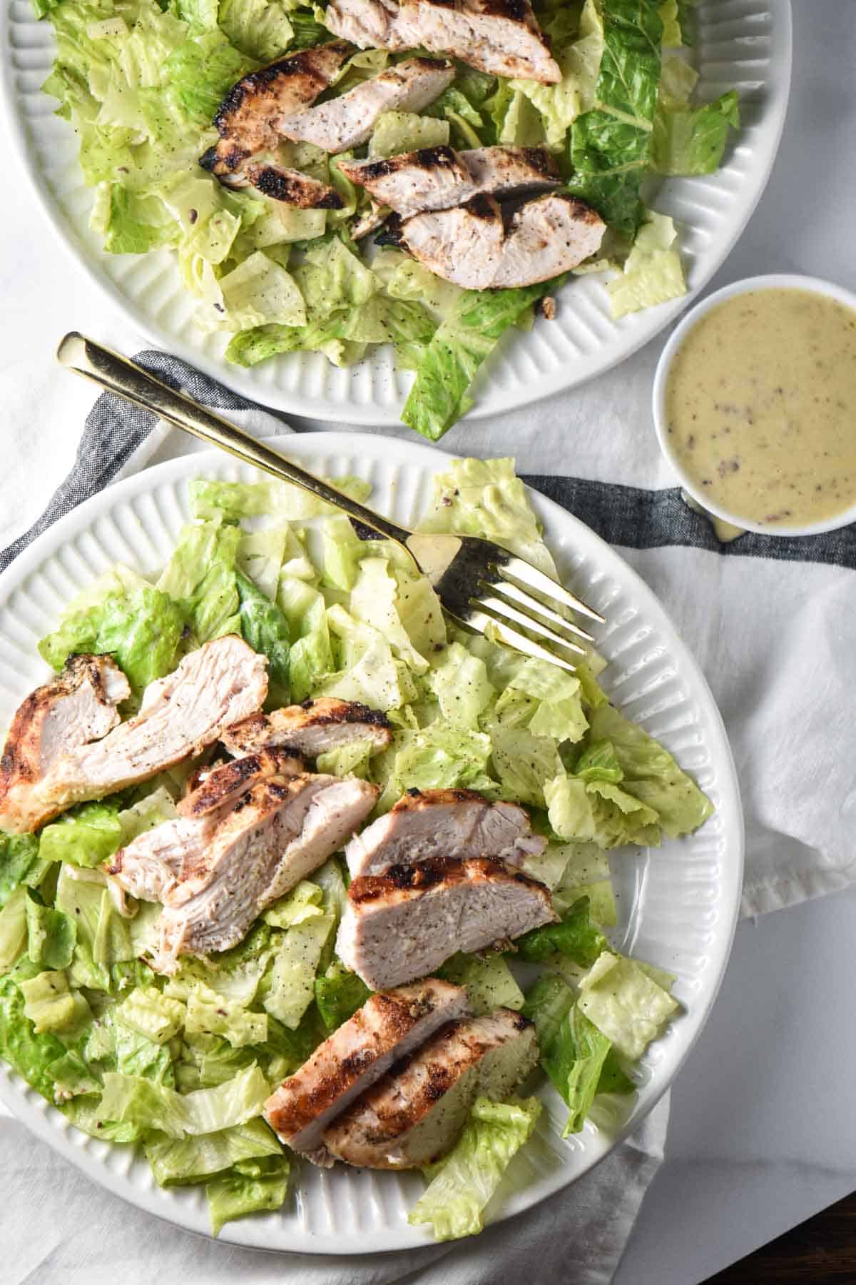 Two Caesar salads next to a bowl of homemade Caesar salad dressing.