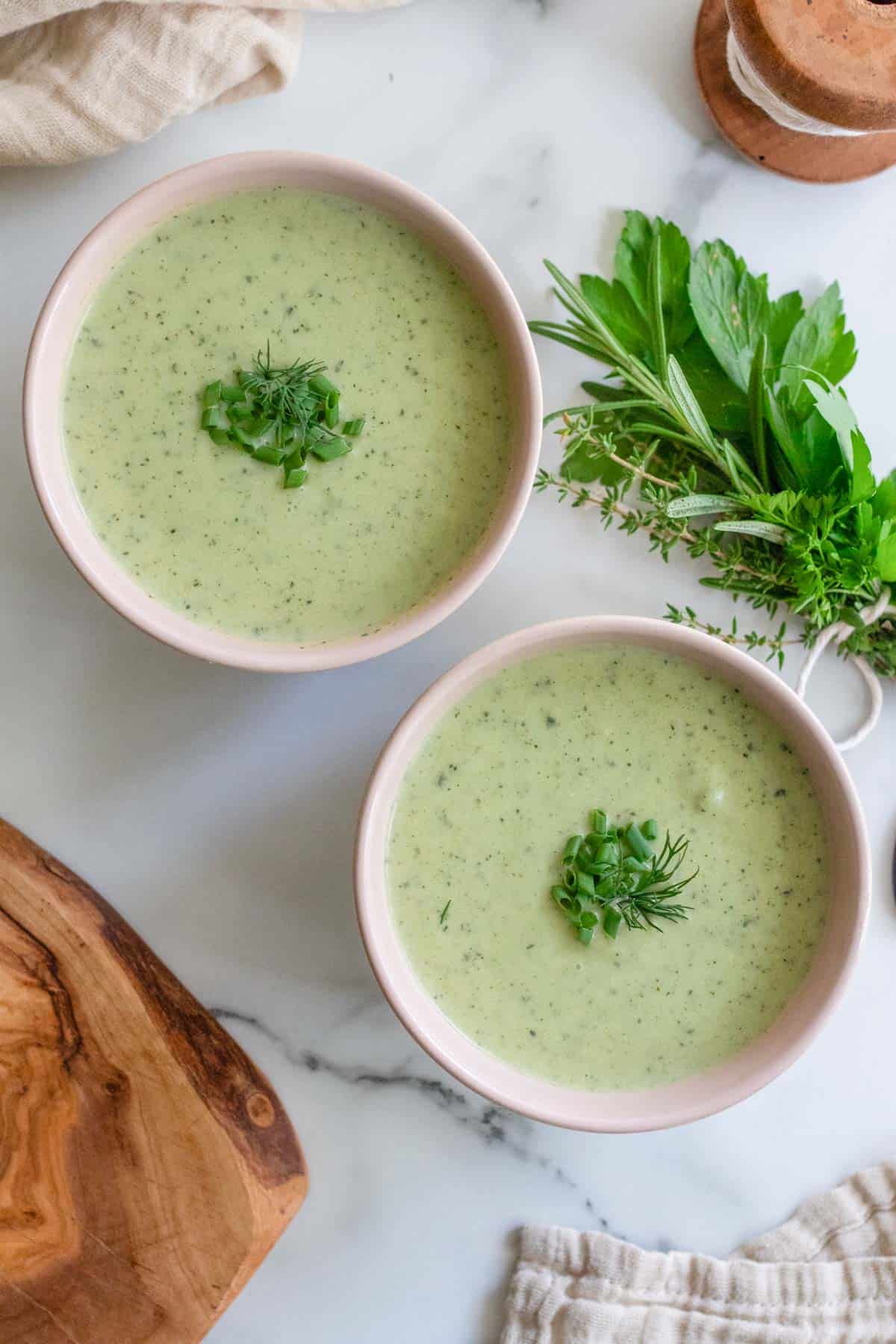 Two pink bowls filled with creamy green soup next to a bunch of fresh parsley and rosemary.