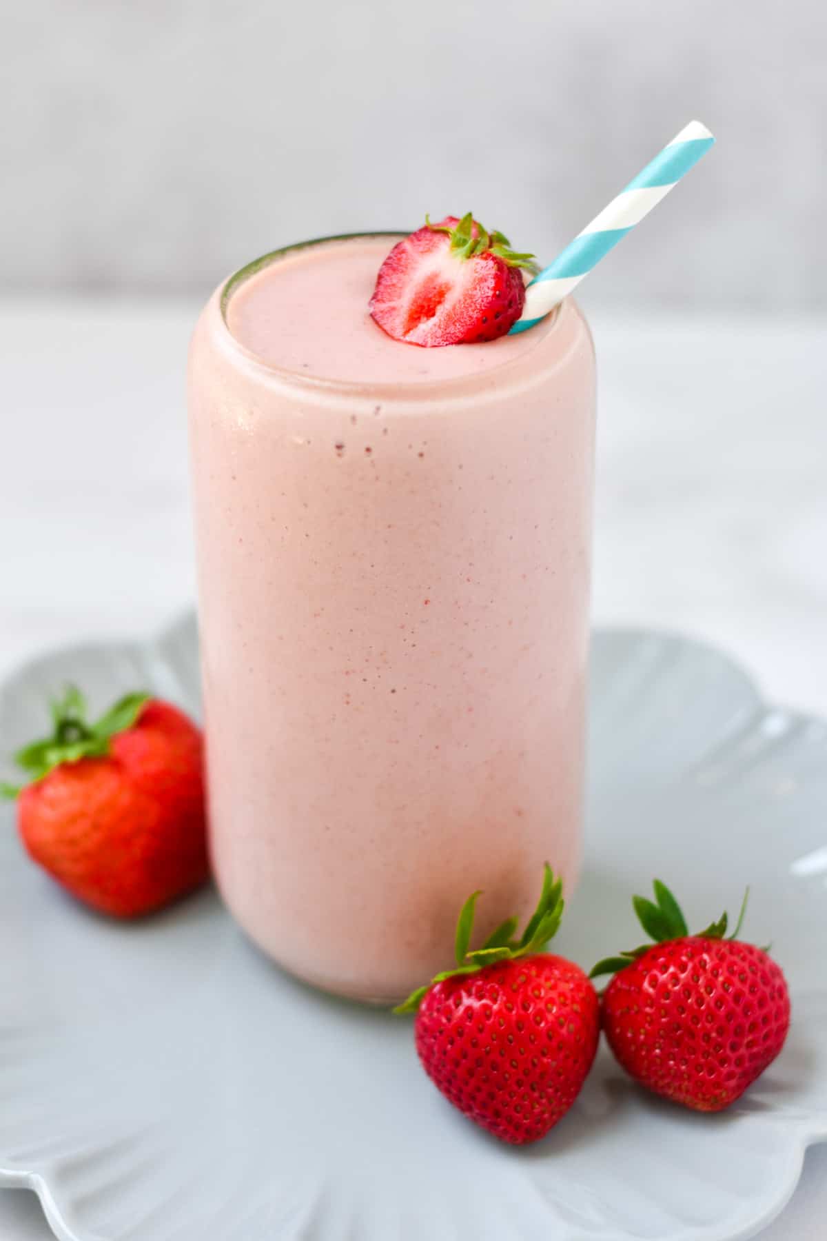 A pink smoothie with a striped straw on a blue plate with strawberries.