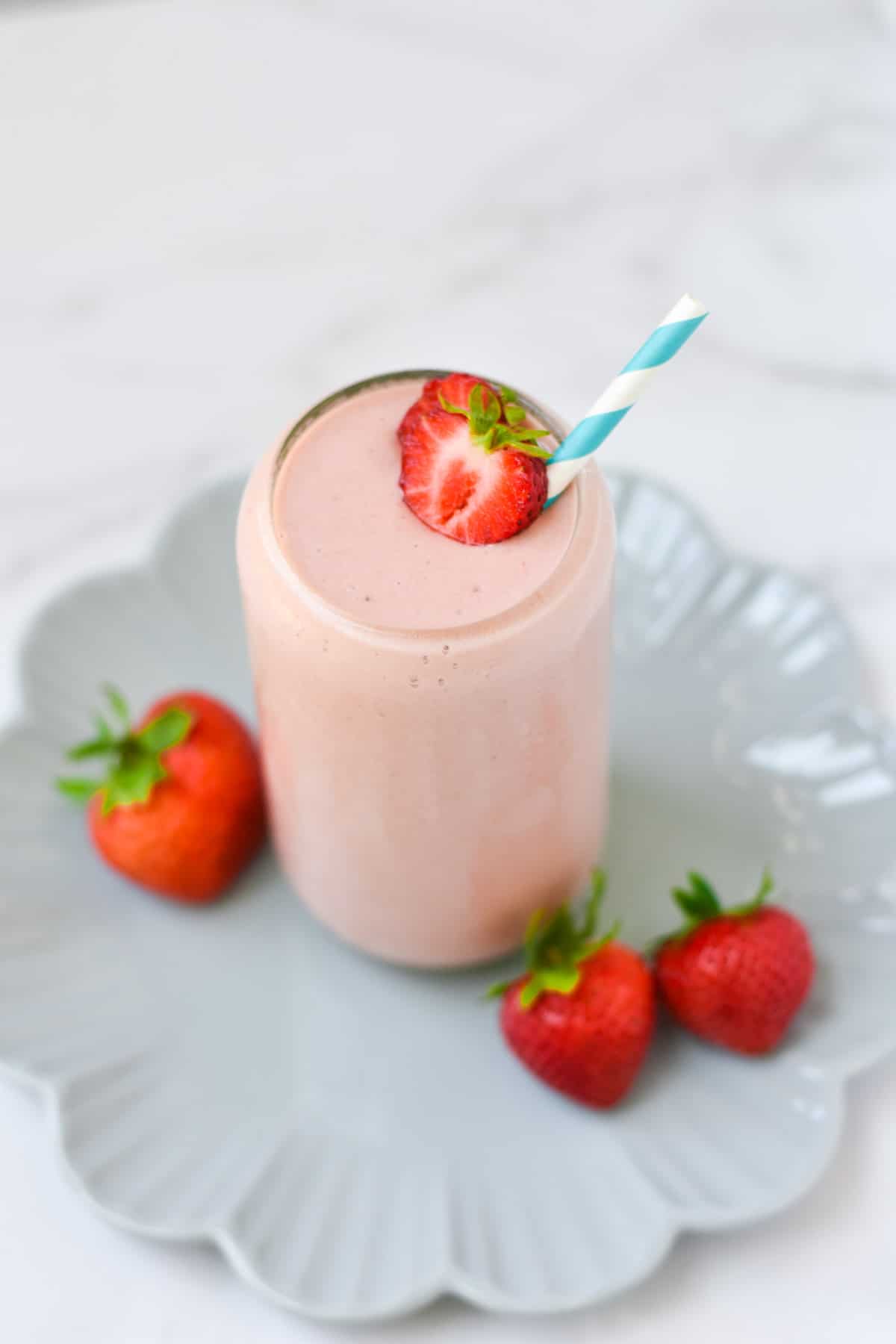 Strawberries on top of a pink smoothie with a blue and white straw and a marble table.