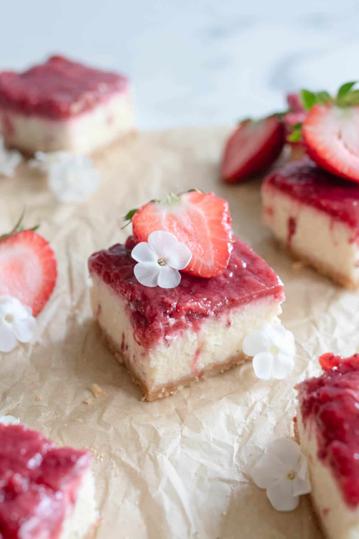 A cheesecake bar topped with strawberries and a white flower on top of brown parchment paper.