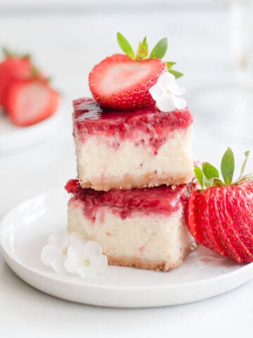 Two strawberry cheesecake bars stacked next to two fresh strawberries and little white flours on a white plate.
