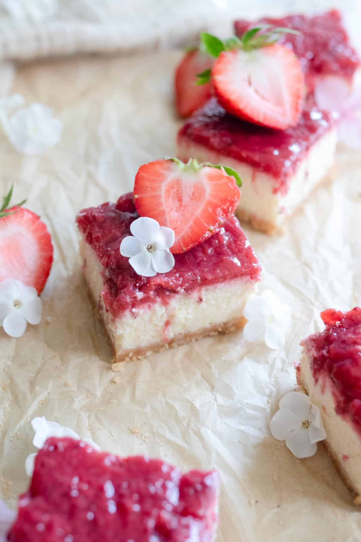 Cut cheesecake bars topped with fresh strawberries and small white flours on brown parchment paper.