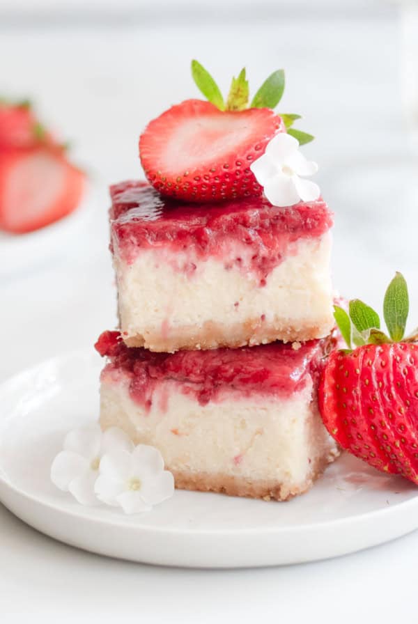 Two strawberry cheesecake bars stacked next to two fresh strawberries and little white flours on a white plate.