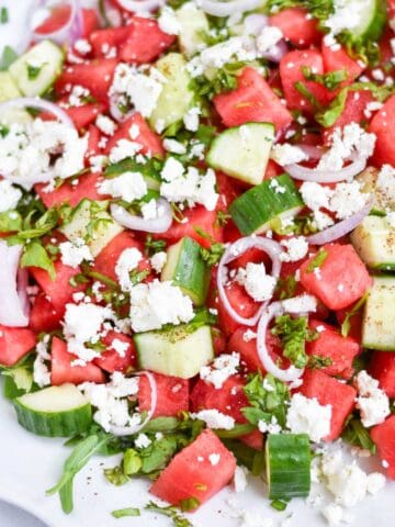 Watermelon, basil, and cucumbers, topped with feta on a white plate.